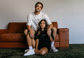 Couple wearing WNGSPN embroidered tees with quote designs, styled together on a leather sofa in a minimal setting