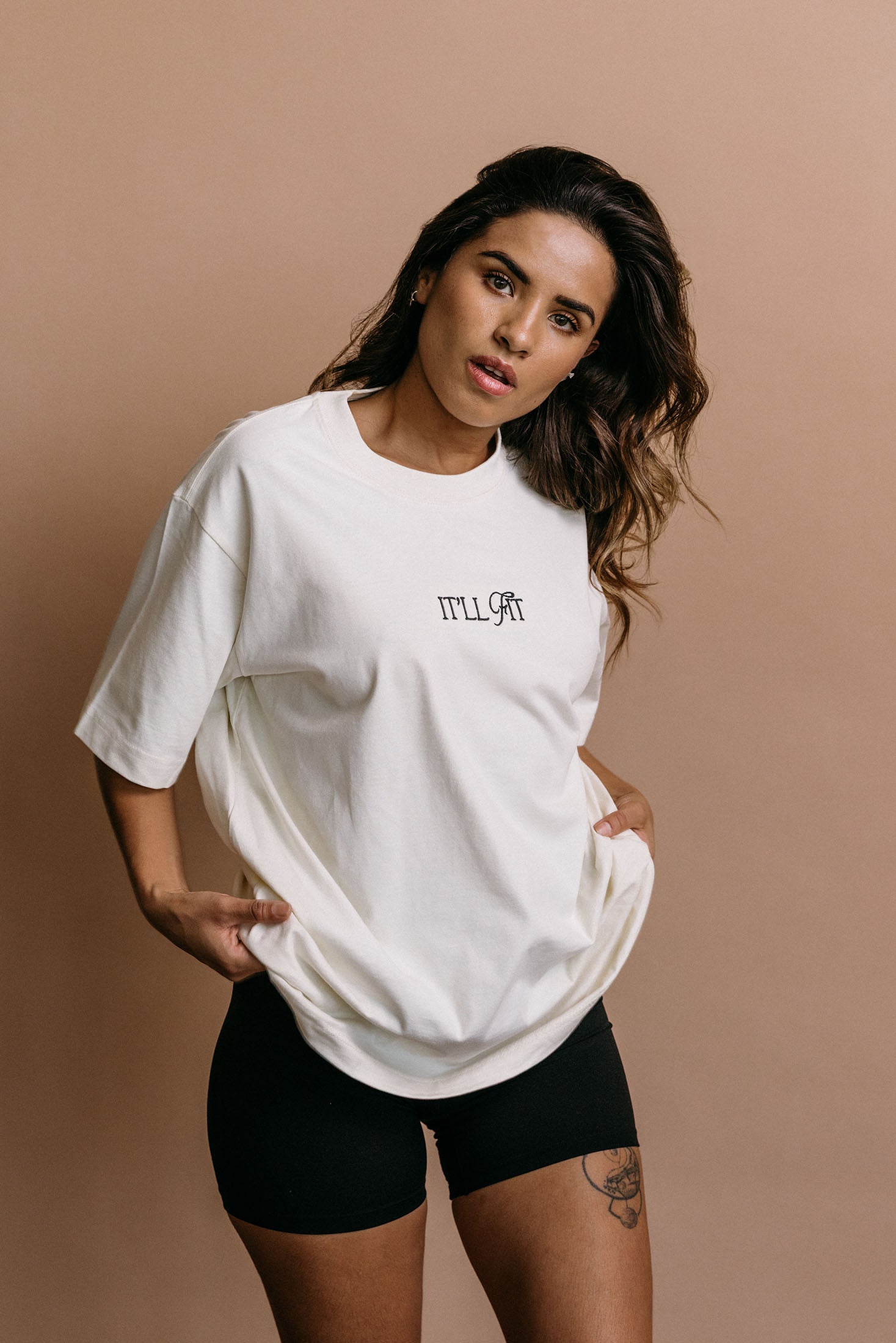 Female model wearing the ecru "It'll Fit" relaxed fit embroidered T-shirt by WNGSPN, styled in a minimal, elevated aesthetic.