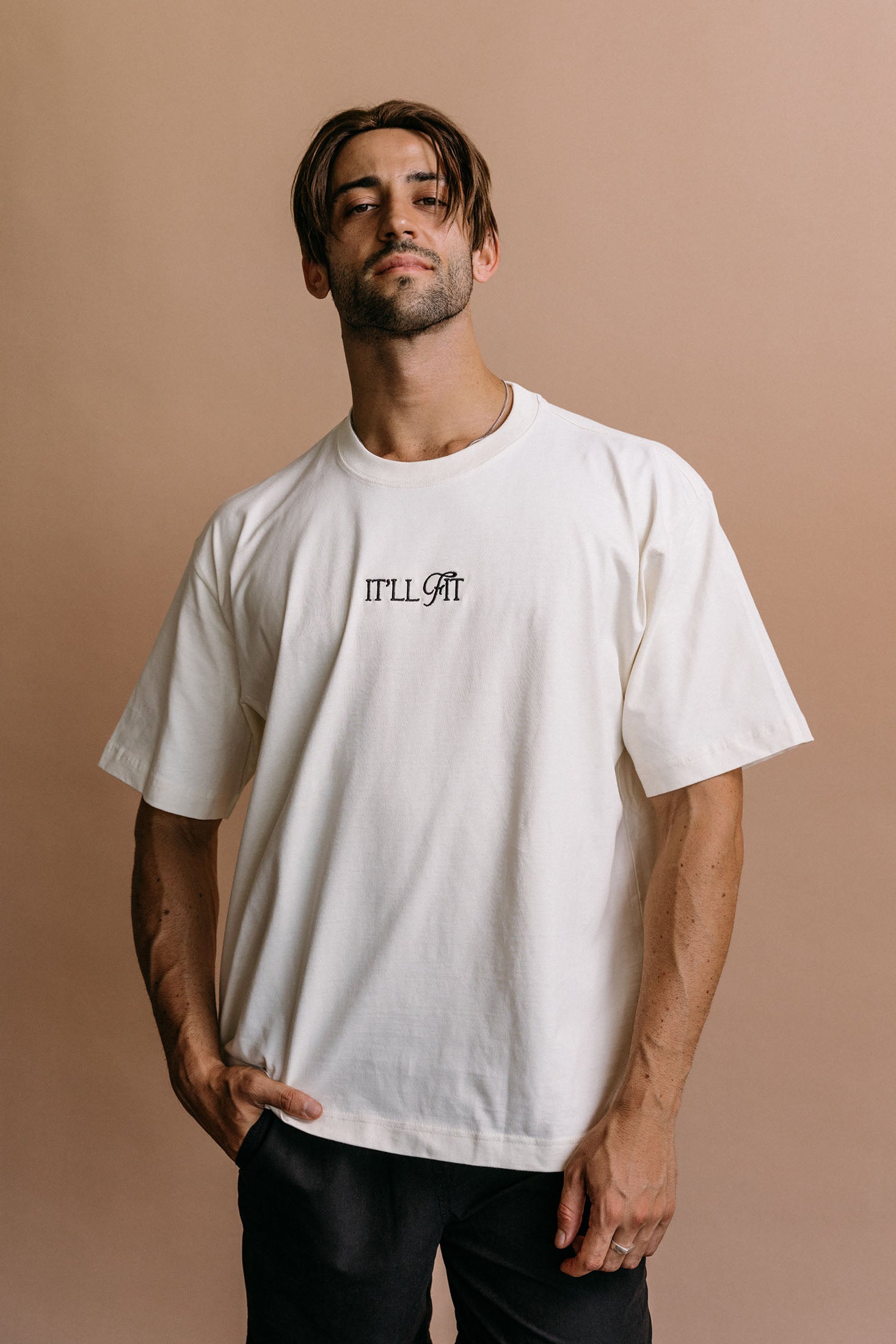 Male model wearing the ecru "It'll Fit" relaxed fit embroidered T-shirt by WNGSPN, styled in a minimal, elevated aesthetic.