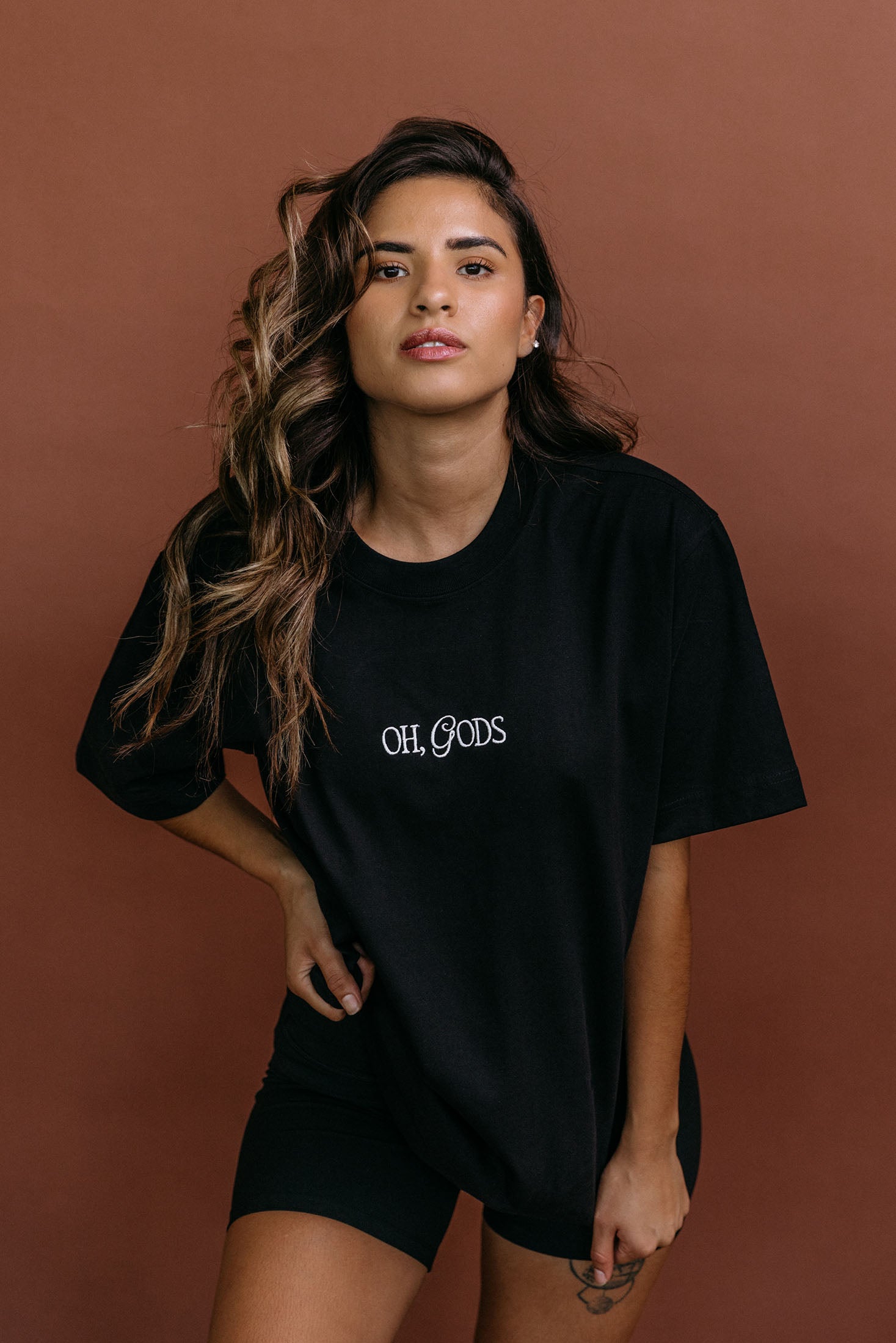 Female model wearing the black "Oh Gods" relaxed fit embroidered T-shirt by WNGSPN, styled with understated confidence.