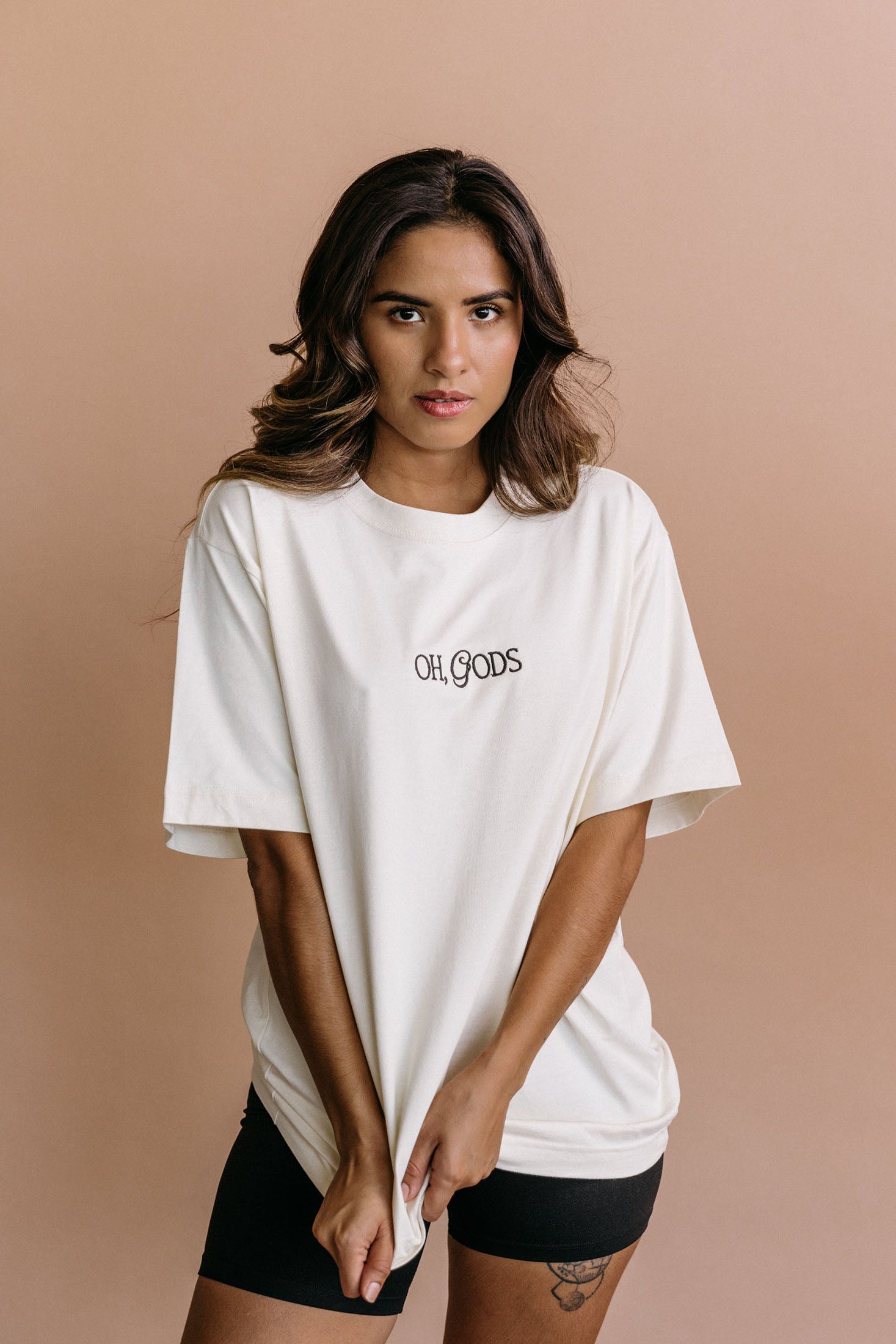 Female model wearing the ecru "Oh Gods" relaxed fit embroidered T-shirt by WNGSPN, styled in a minimal, elevated aesthetic.