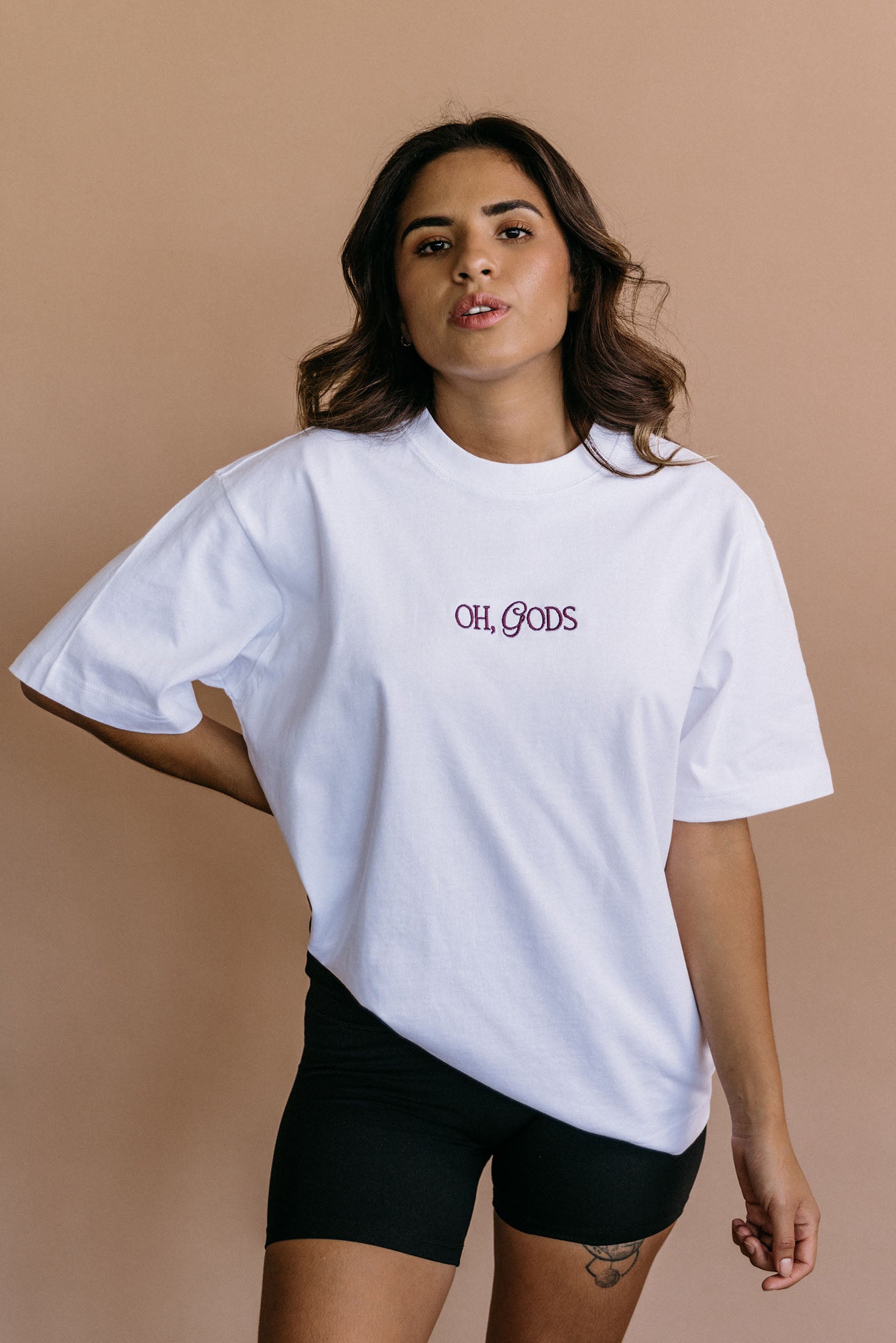 Female model wearing the white "Oh Gods" relaxed fit embroidered T-shirt by WNGSPN, with a relaxed silhouette and clean lines.