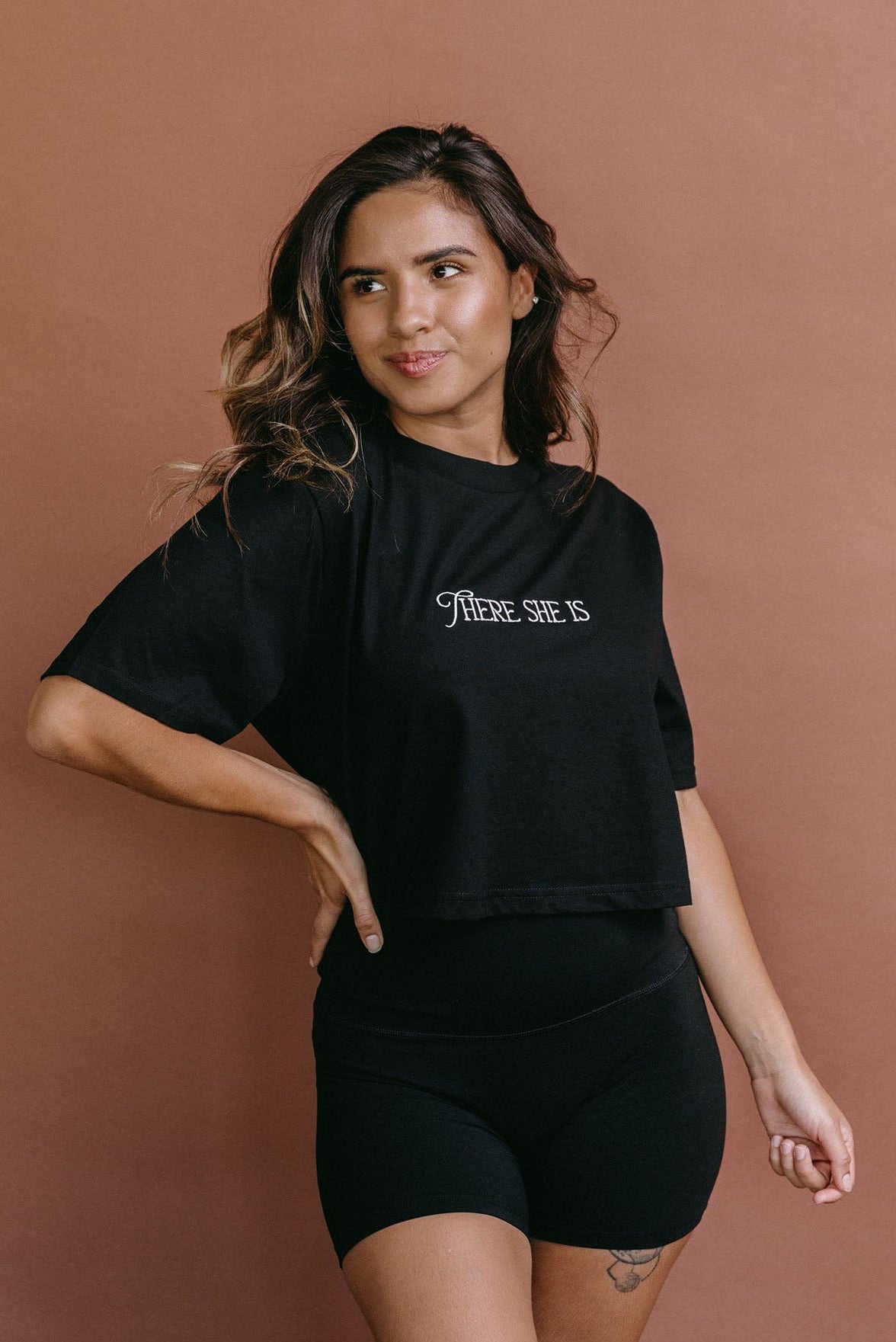 Female model wearing the black "There She Is" cropped embroidered T-shirt by WNGSPN, styled with understated confidence.