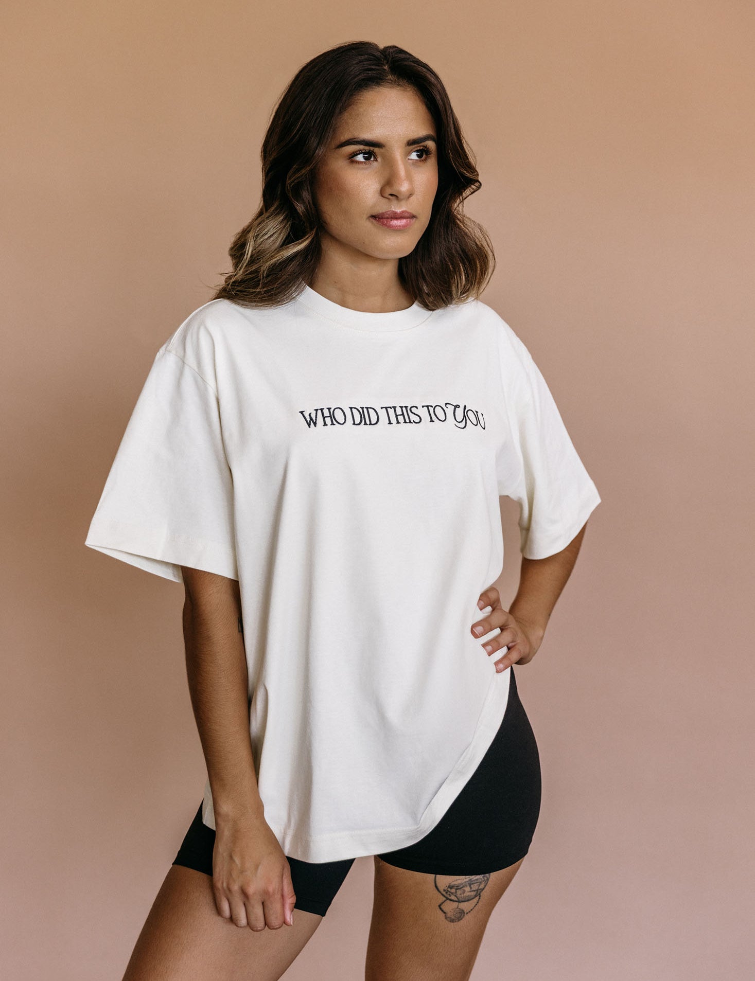Female model wearing the ecru "Who Did This To You" relaxed fit embroidered T-shirt by WNGSPN, styled in a minimal, elevated aesthetic.