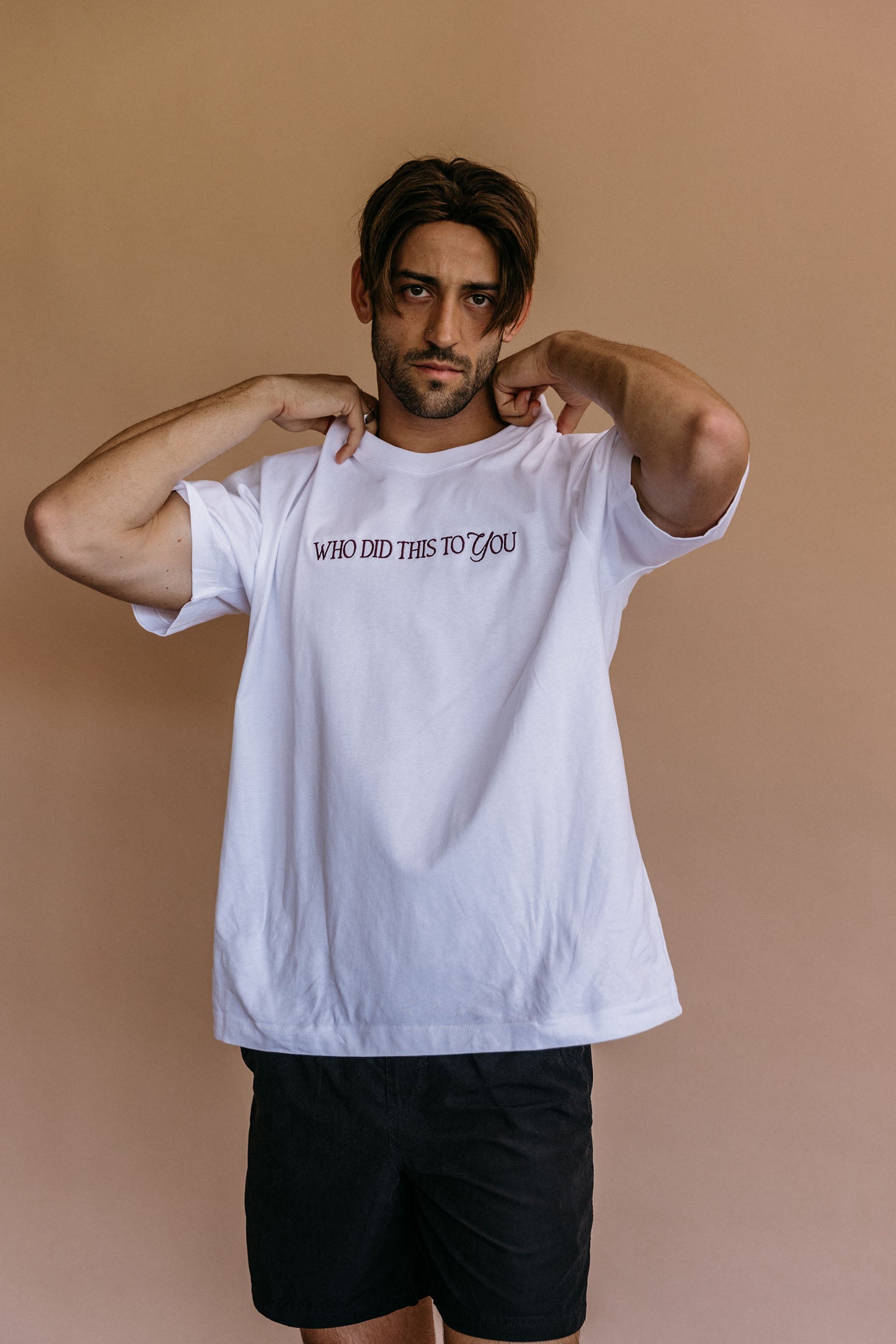 Male model wearing the white "Who Did This To You" relaxed fit embroidered T-shirt by WNGSPN, with a relaxed silhouette and clean lines.